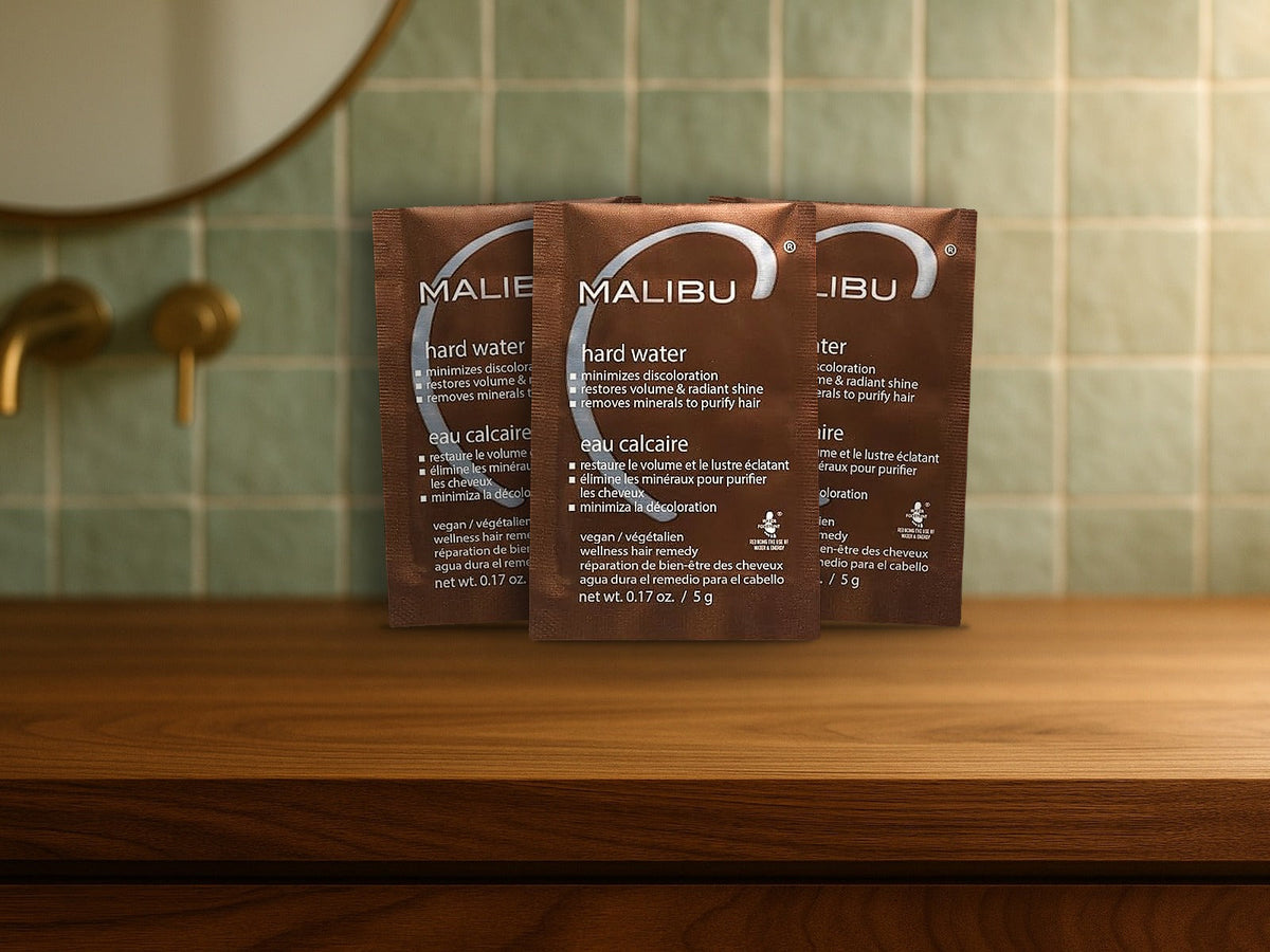 Malibu C Hard Water Wellness Remedy packets standing on a walnut bathroom vanity in front of sage green tile, product clearly in focus