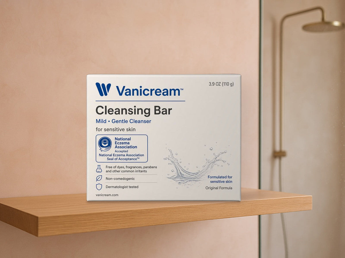Vanicream Cleansing Bar box standing on a light oak floating shelf against a blush pink plaster wall, with a glass shower panel and a brass rain shower head softly blurred at right