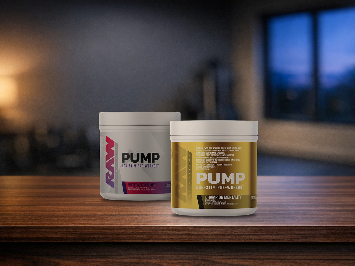 Two tubs of RAW Pump Non-Stim Pre-Workout, a gold Champion Mentality tub in front and a white Watermelon Grape tub behind it, on a dark walnut wood top in a dimly lit room with a warm lamp glow at left and a window showing blue evening sky at right