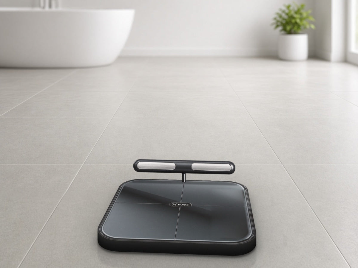 Hume Body Pod smart scale with its handheld sensor bar docked, lying on a light grey tiled bathroom floor near a freestanding tub and a potted plant
