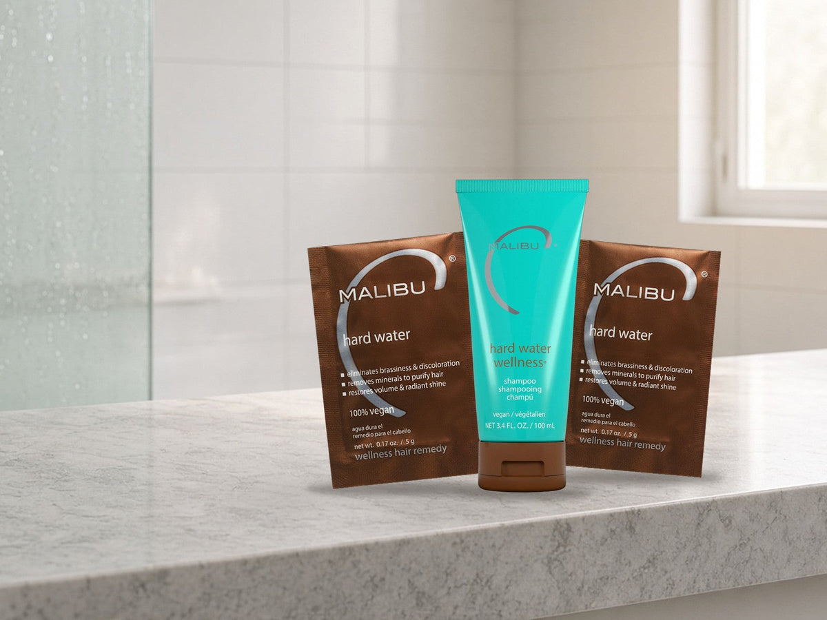 Malibu C Hard Water Wellness Shampoo tube standing between two Malibu C Hard Water Wellness Remedy packets on a light marble bathroom ledge in soft morning light, products clearly in focus