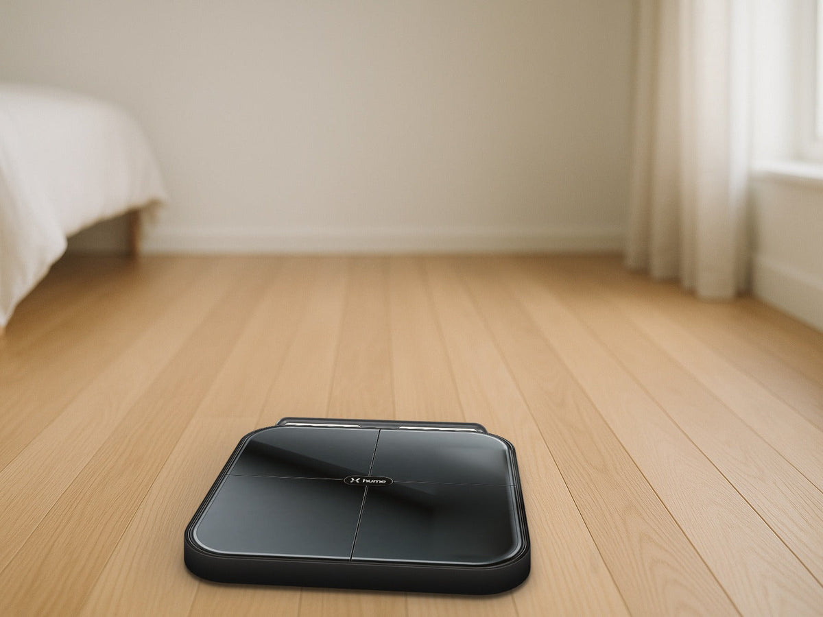 Hume Health Body Pod smart scale with its handlebar docked flush at the back on a light oak floor in a bright bedroom with soft morning window light, product clearly in focus