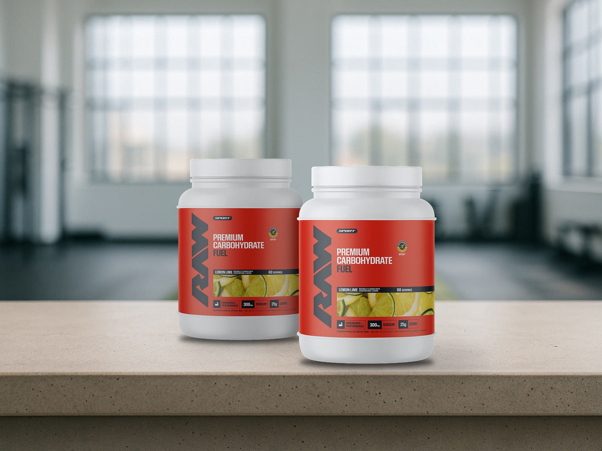 Two tubs of RAW Fuel Carbohydrate Drink Mix Lemon Lime on a pale concrete ledge in a bright gym, product clearly in focus