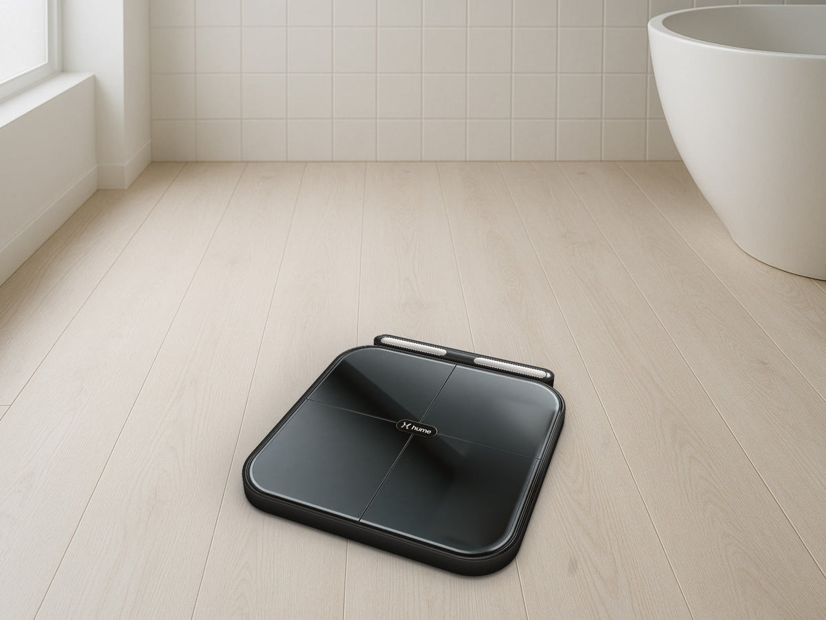 Black Hume Body Pod smart scale with its handlebar docked along the back edge, on a pale wood floor in a bright bathroom with white tile walls and a white freestanding bathtub