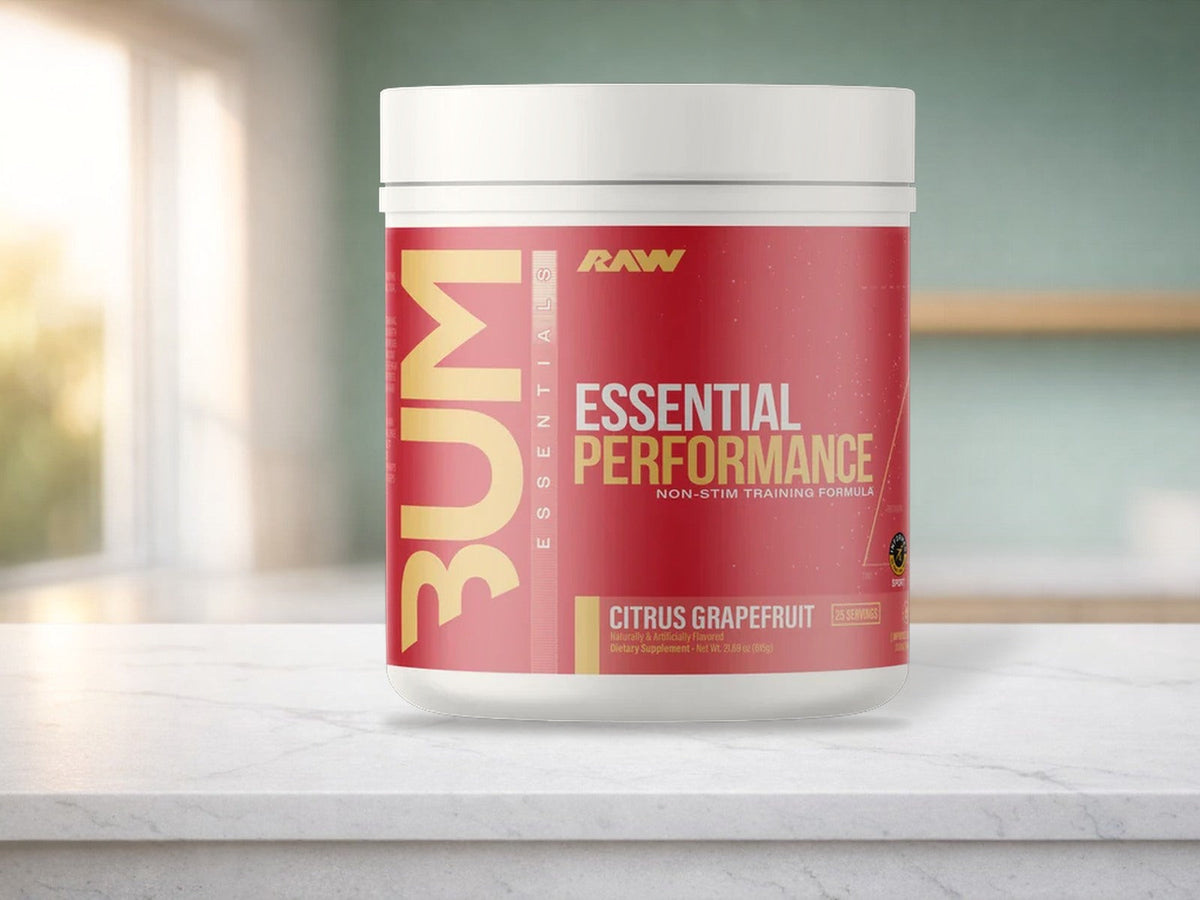 A red RAW Essential Performance Citrus Grapefruit pre-workout tub with a white lid standing on a white stone kitchen counter, with a sage green wall, an empty wooden shelf, and a sunny window softly blurred behind it