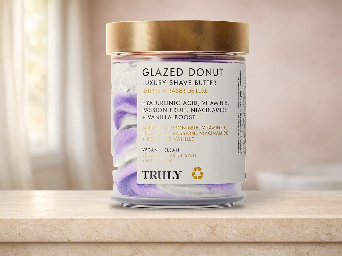 A glass jar of Truly Glazed Donut Luxury Shave Butter with a gold lid and swirled purple and white butter, standing on a cream travertine bathtub ledge in front of a blush pink wall and a window with sheer curtains